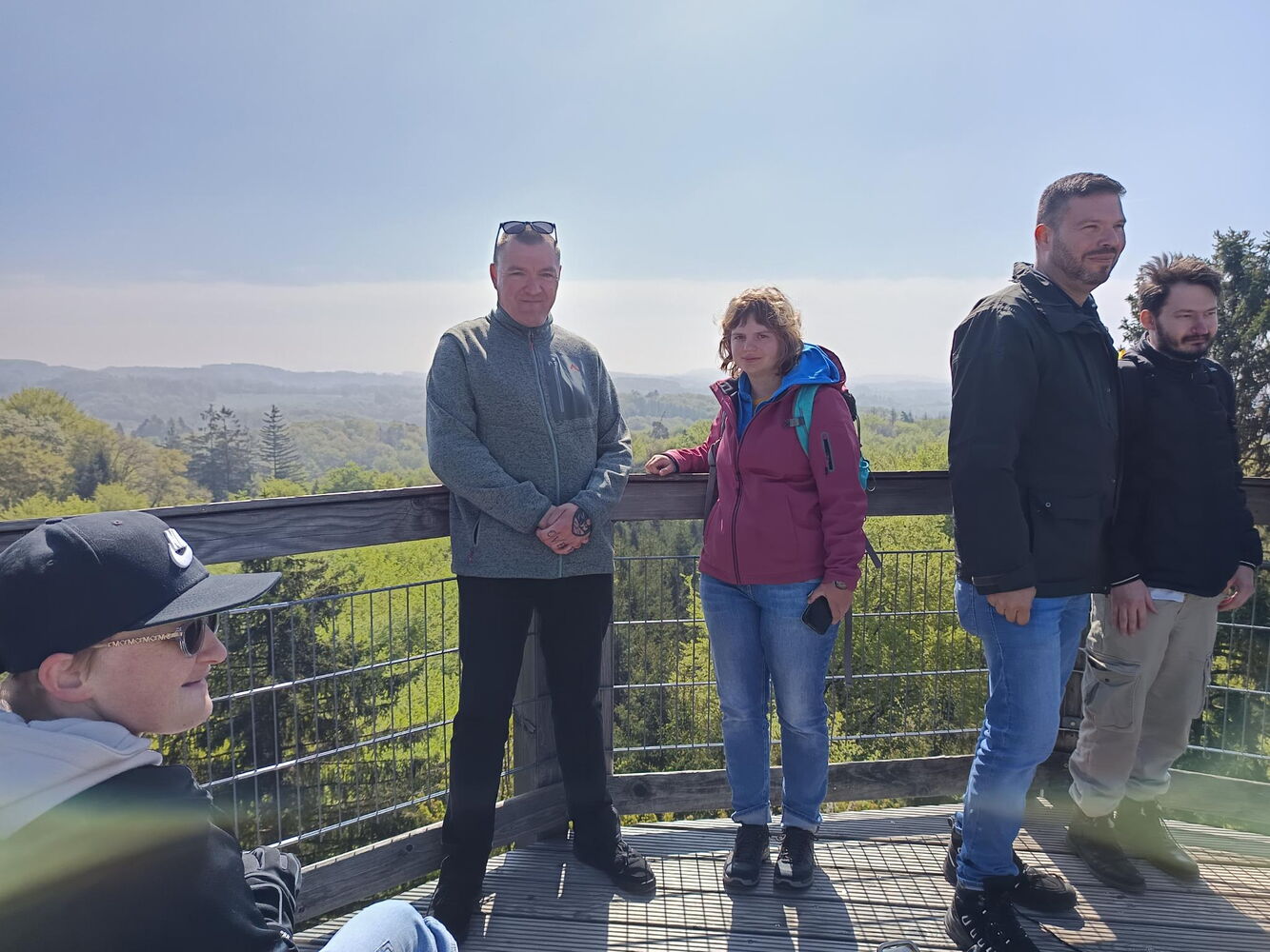 Vor einer Brüstung aus Metallgitter und Holz stehen fünf Personen. Im Hintergrund sind grüne Baumwipfel zu sehen.