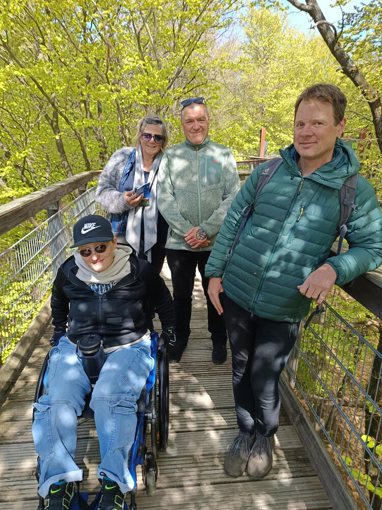 Auf einer Brücke, dem Baumwipfelpfad, stehen vier Personen, eine davon im Rollstuhl. Die Sonne scheint, drei Personen tragen Sonnenbrillen. Im Hintergrund sind grüne Baumwipfel zu sehen.