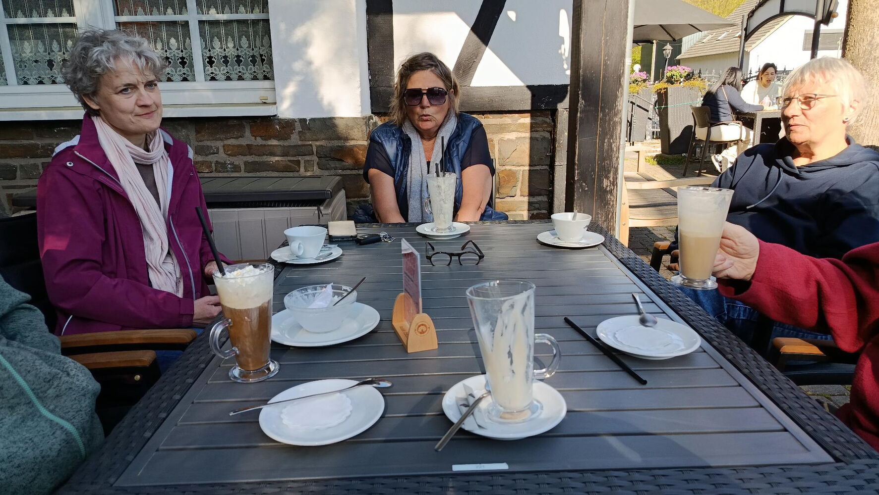 Eine Gruppe sitzt an einem Tisch, mit Kaffee-Getränken vor sich. Im Hintergrund ist ein Fachwerkhaus.