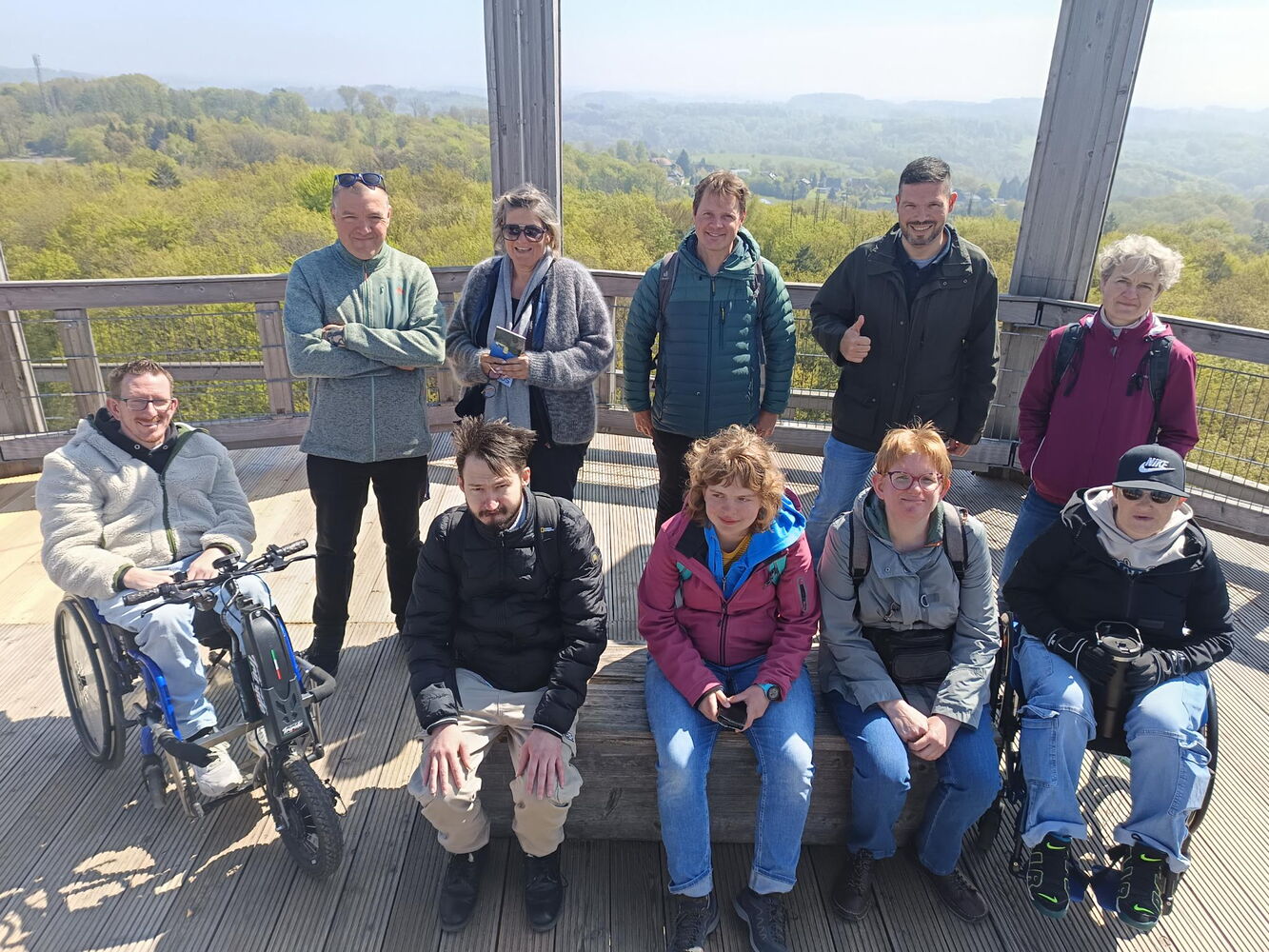 Zehn Personen, teilweise stehend, teilweise sitzend oder im Rollstuhl, befinden sich auf dem Aussichtsturm und posieren für ein Gruppenbild. Im Hintergrund sind grüne Baumwipfel zu sehen, man kann sehr weit in die Distanz sehen. Es ist sonnig.