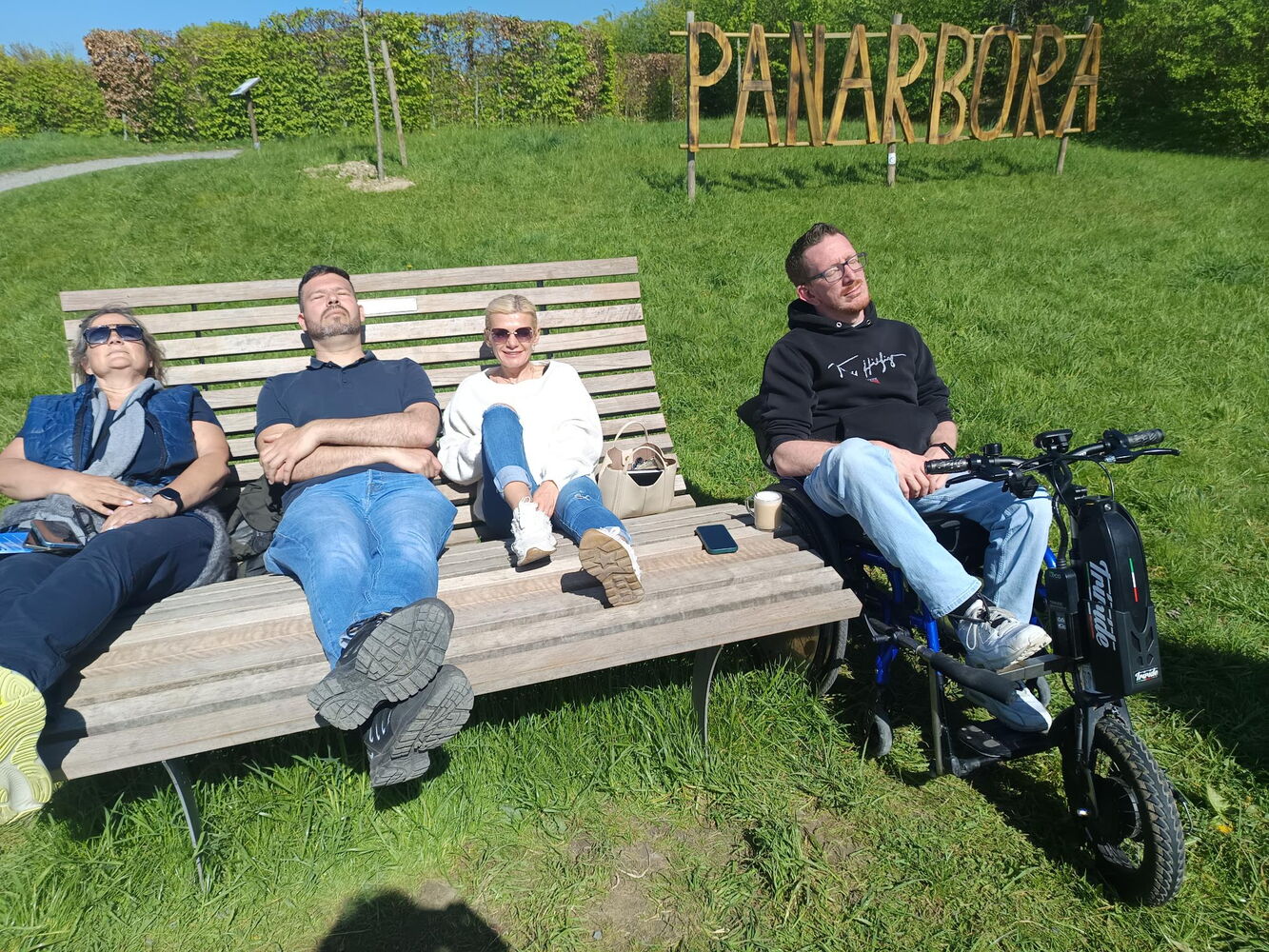 Auf einer grünen Wiese ist ein Holzaufsteller mit dem Namen Panarbora. Im Vordergrund ist eine Sonnenliege aus Holz, auf der drei Personen liegen. Neben der Bank sitzt eine weitere Person im Rollstuhl. Die Augen sind geschlossen und sie genießen die Sonne